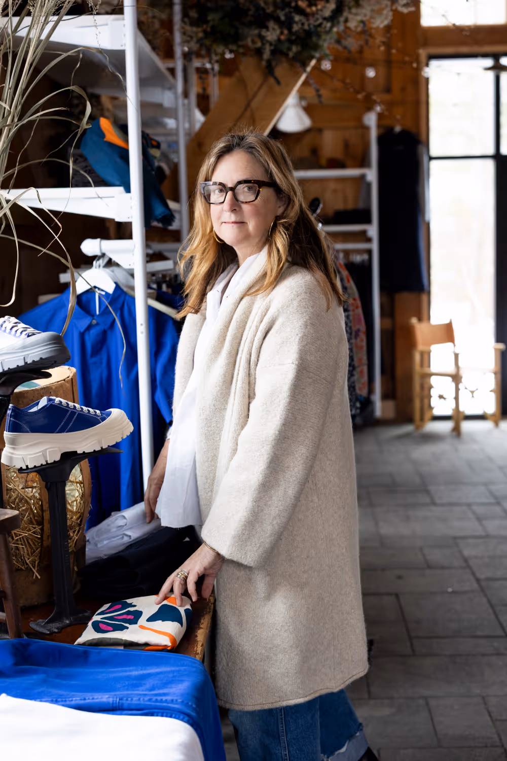 Woman wearing glasses and a beige coat in a boutique with clothing and shoes displayed.