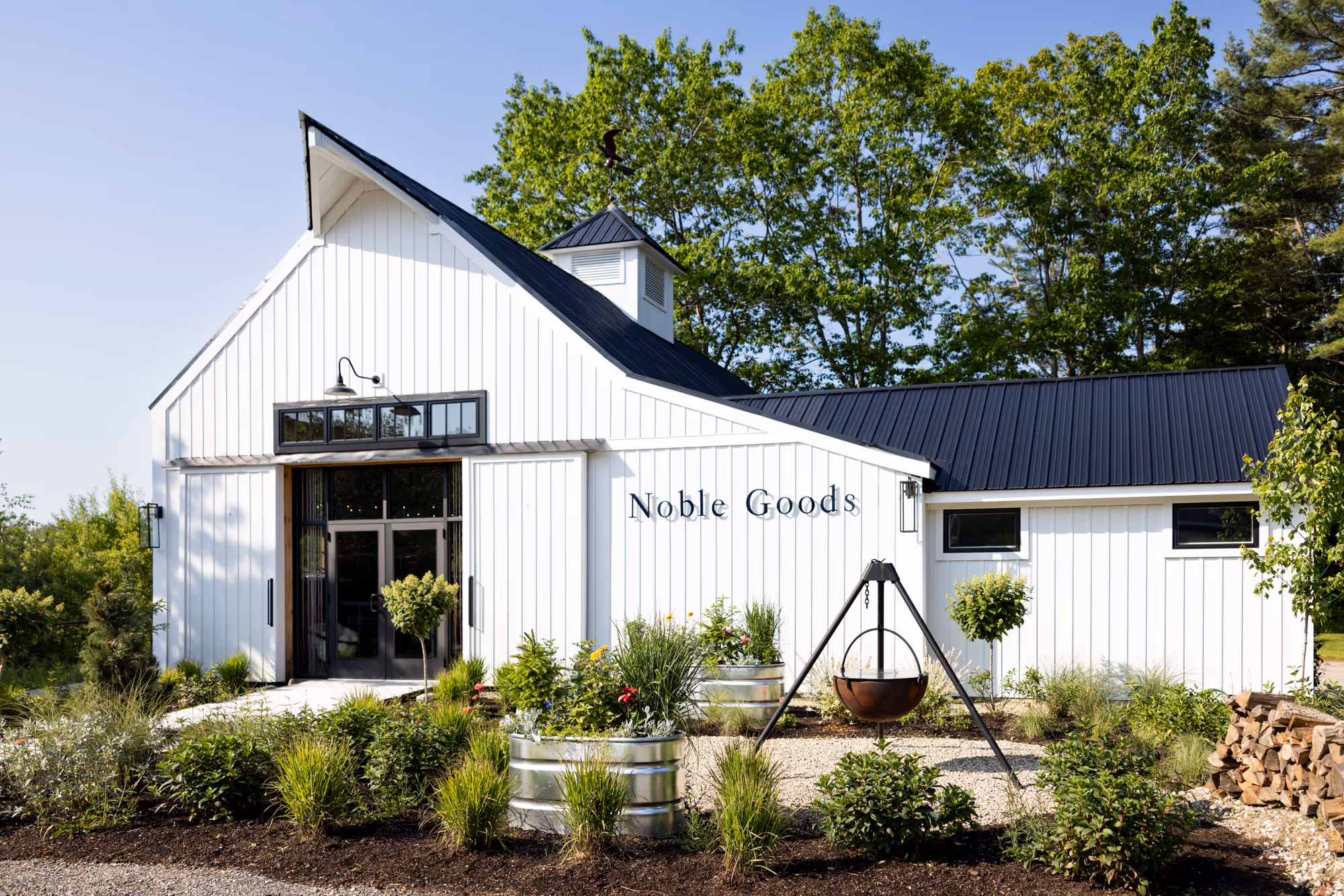 White barn-style building with black roof, entrance doors, garden beds, and a fire pit in front.
