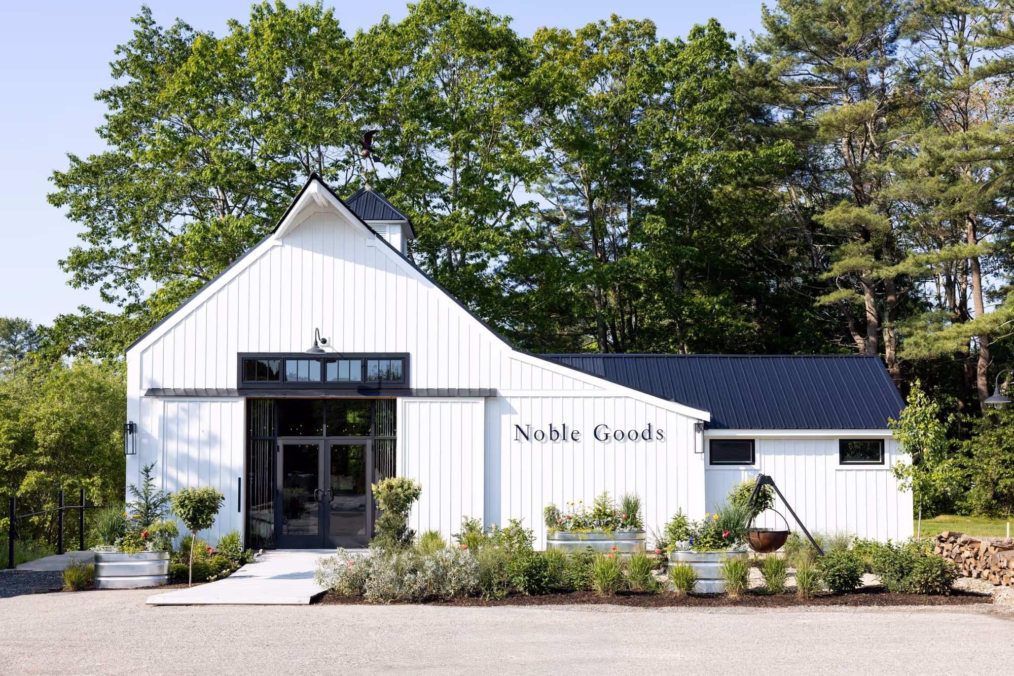 White building with black roof and glass doors labeled 'Noble Goods' surrounded by green trees and plants.