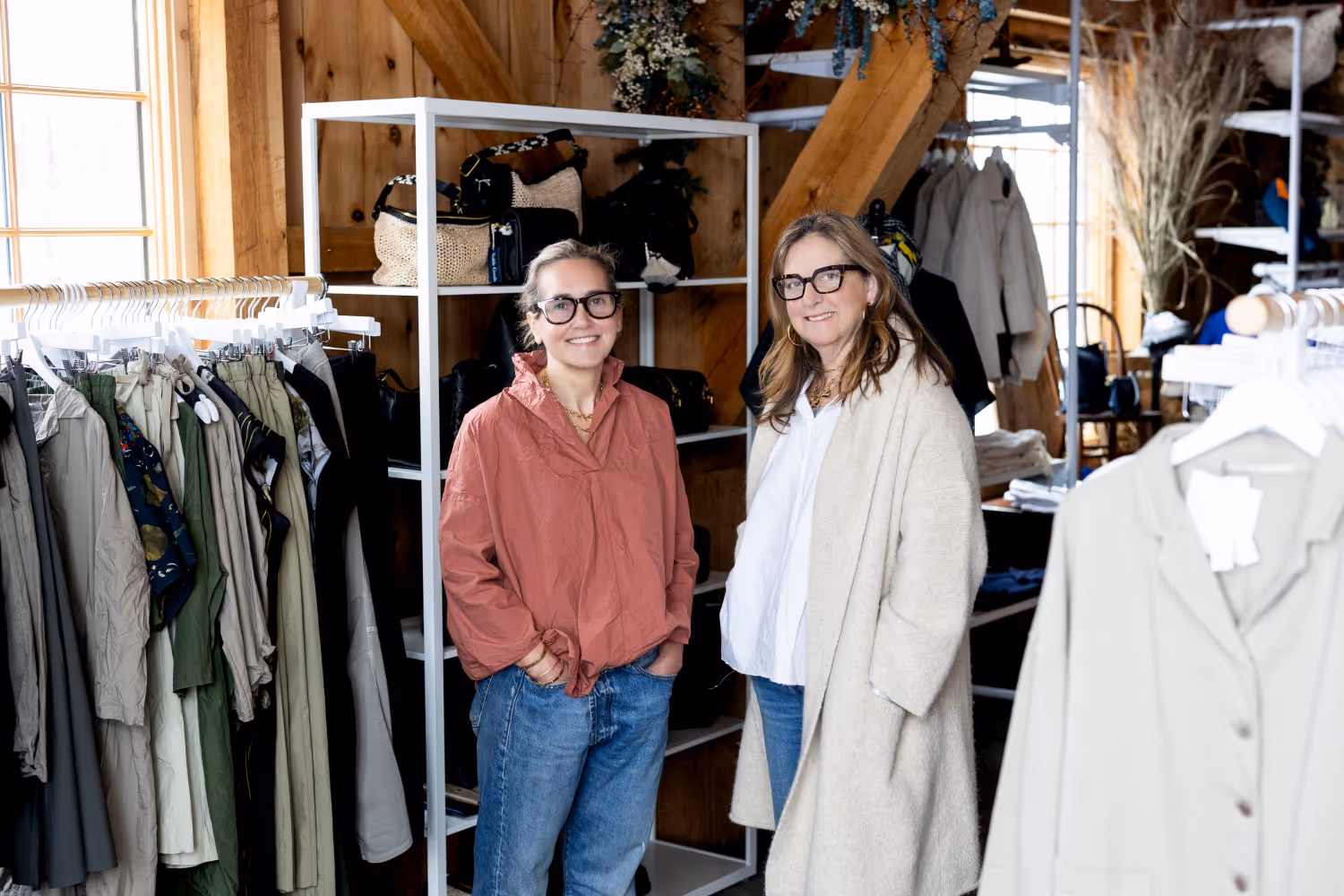Two smiling women wearing glasses standing in a clothing store with racks of clothes and purses.