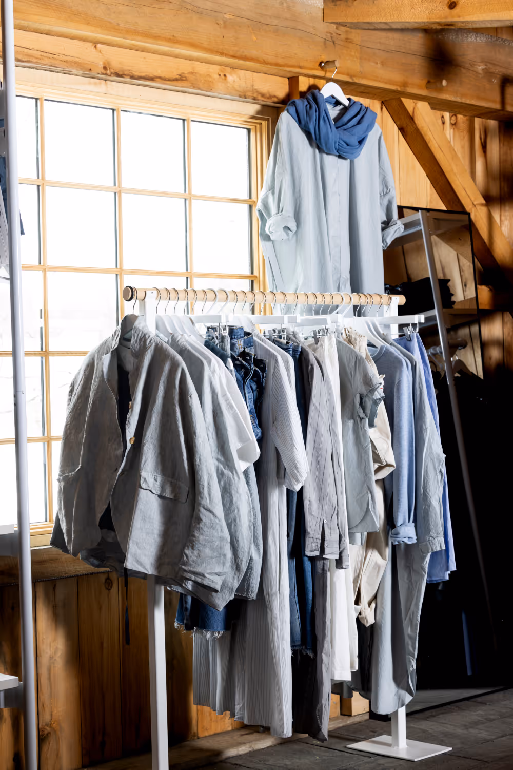 Clothing rack with various light-colored shirts and pants in a wooden room with a window.