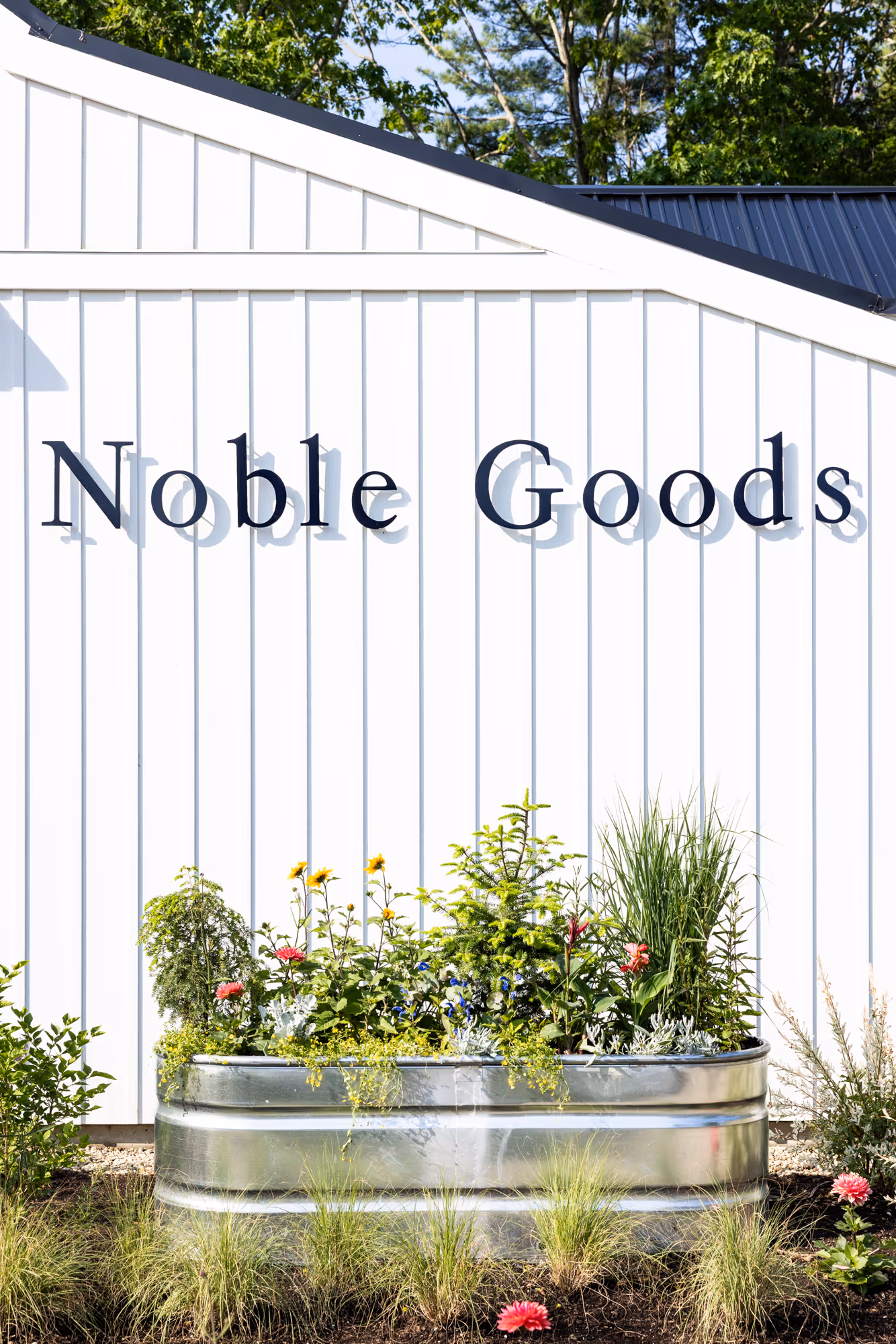 Raised metal planter with flowers and plants in front of white building with Noble Goods sign.