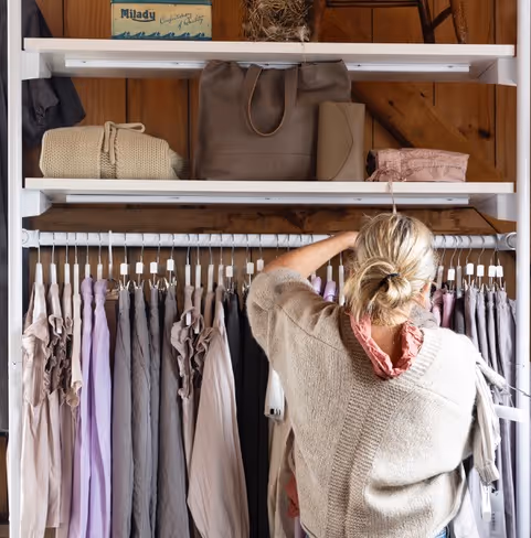Person browsing beige, lavender, and gray clothes on white rack in a wooden-paneled room.