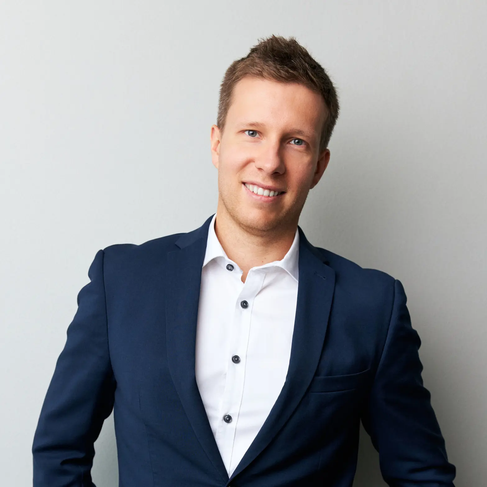 Smiling man in a navy blazer and white shirt standing against a light gray wall.