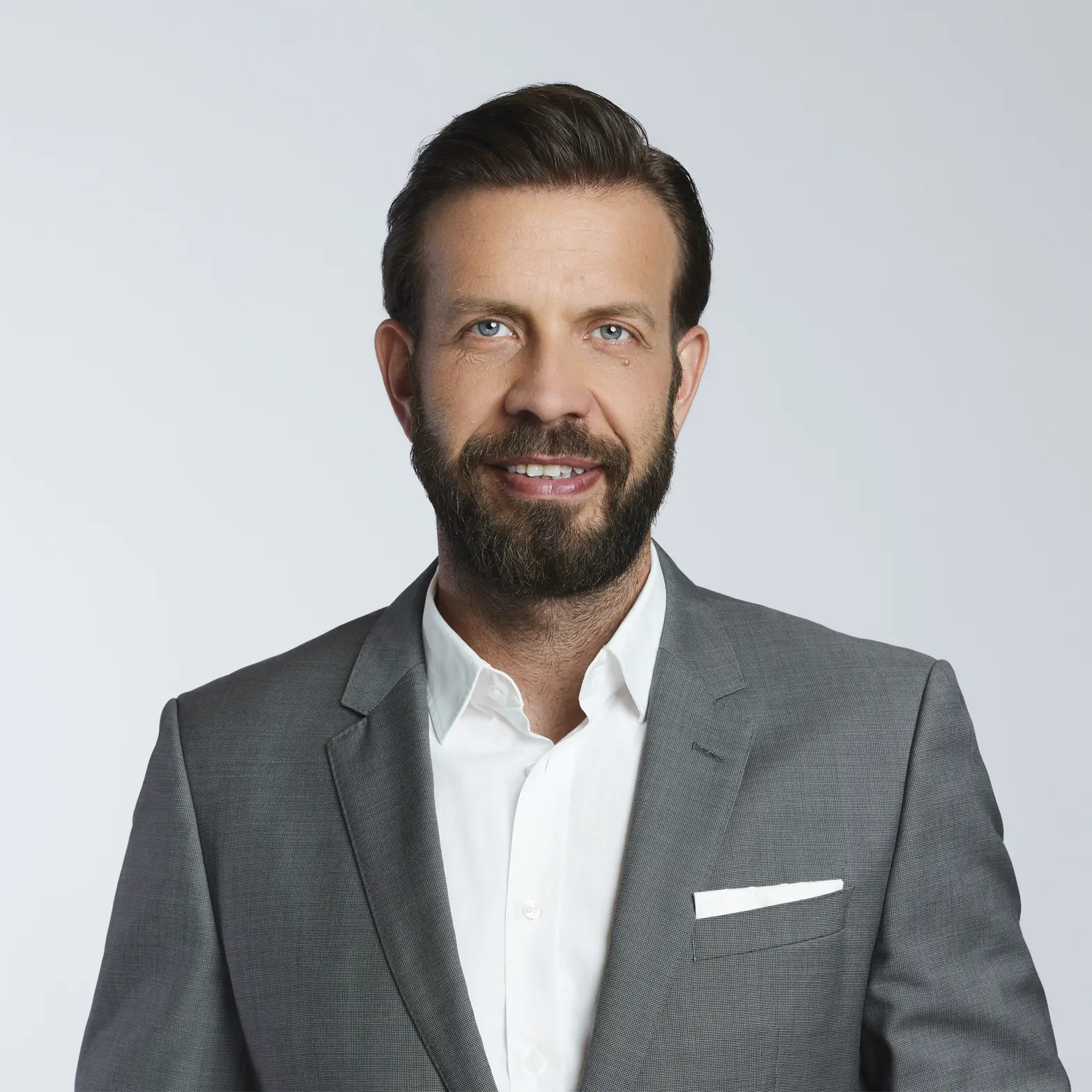 Smiling man with beard wearing a grey suit jacket and white shirt against a plain background.