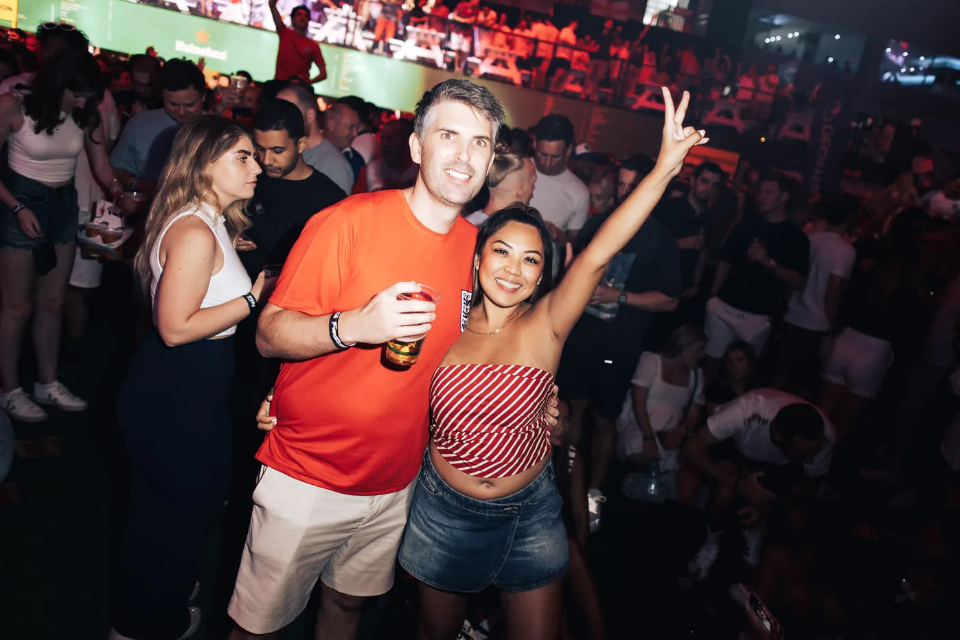 Smiling man in orange shirt with drink and woman in striped top raising arm at crowded party.