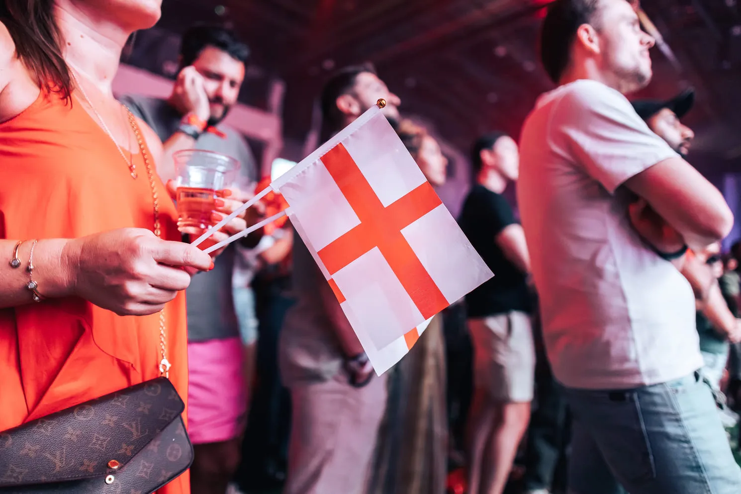 Person in orange dress holding two small England flags in a crowd of people at an event.