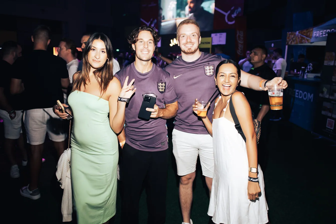 Four friends smiling at a party, two men in matching shirts, women in green and white dresses, holding drinks.