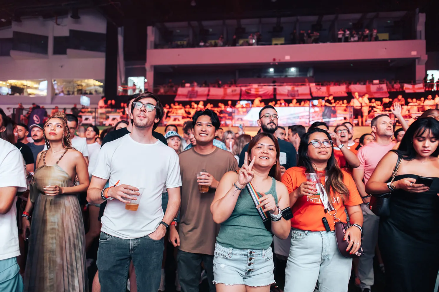 Diverse crowd at an indoor event, some holding drinks, one woman making a peace sign.