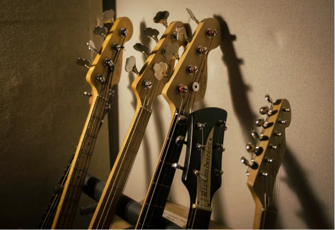 Close-up of five guitar and bass headstocks with tuning pegs against a wall background.