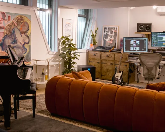 Modern music studio with piano, guitar, orange sofa, desk with computer monitors, and artwork.