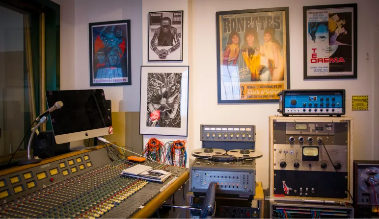 Music studio with mixing desk, microphone, reel-to-reel tape recorder, and framed posters on the wall.