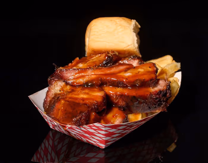 Barbecue ribs coated in sauce served with a dinner roll and potato chips in a red checkered tray.
