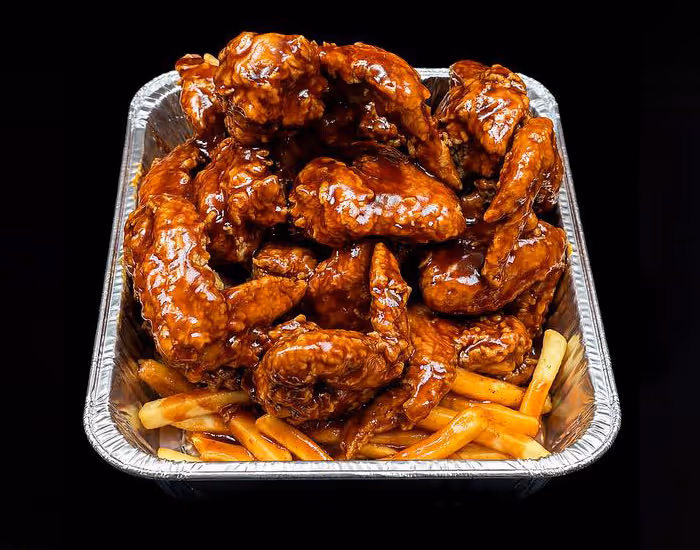 Aluminum tray filled with barbecue-glazed chicken wings atop golden French fries against a black background.