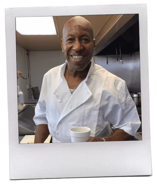 Smiling male chef in white uniform holding a white cup in a commercial kitchen.