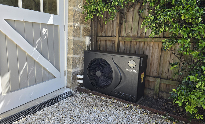 Black Austek outdoor heat pump unit installed beside a stone wall and wooden fence surrounded by gravel and greenery.