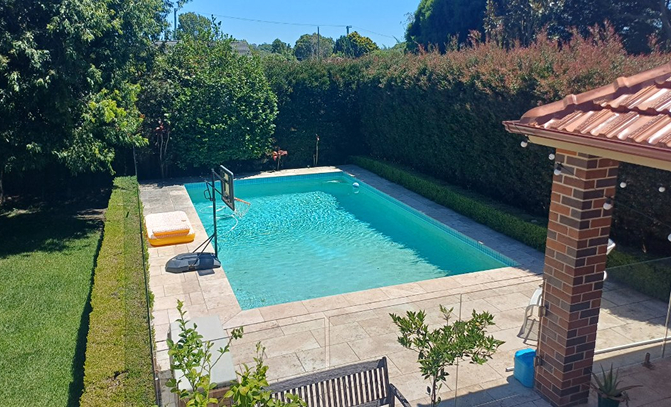 Rectangular swimming pool with clear blue water in a backyard surrounded by green hedges and trees.