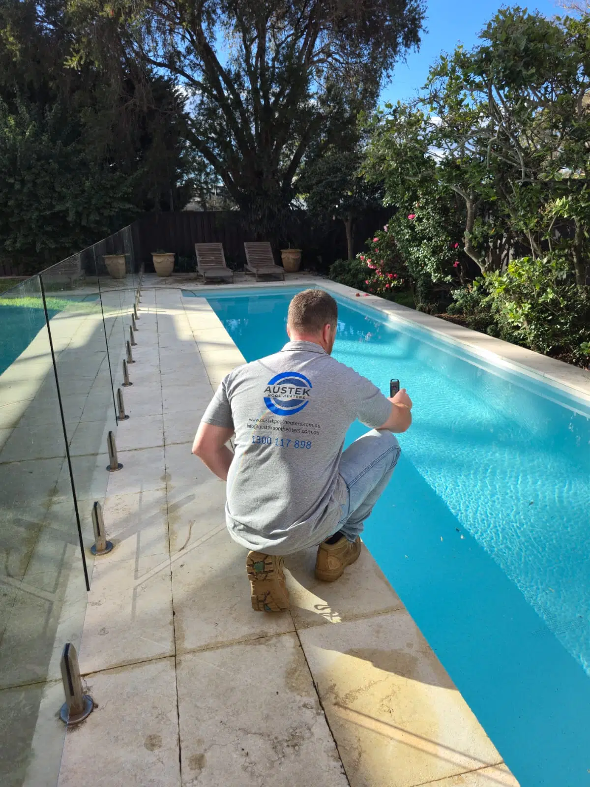 Technician in gray shirt kneeling by a clear glass fenced pool with blue water in a sunny backyard.