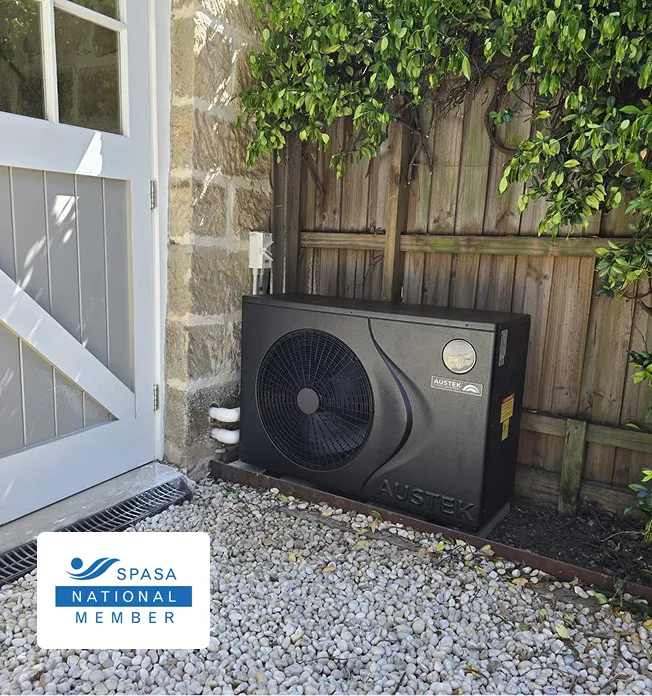 Austek pool heater unit installed beside a house wall and wooden fence on gravel ground.