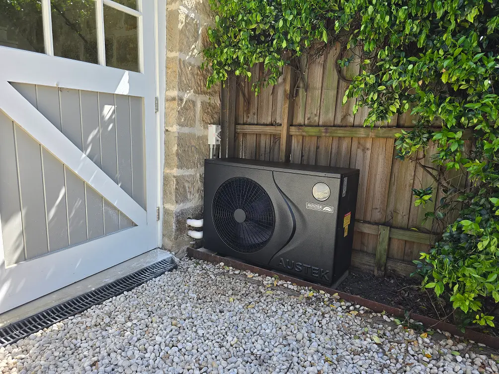 Black Austek pool heat pump installed next to a stone wall and wooden fence on gravel ground.