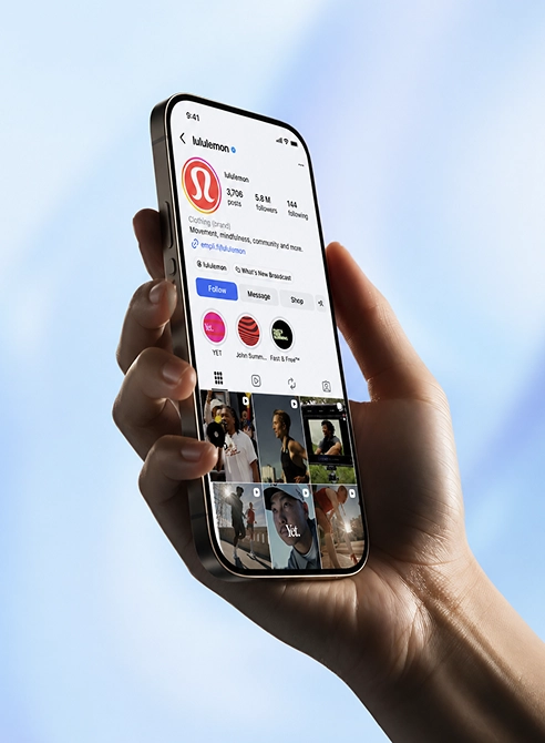 Hand holding a smartphone displaying Lululemon's verified Instagram profile showing posts, followers, and story highlights against a blue sky background.