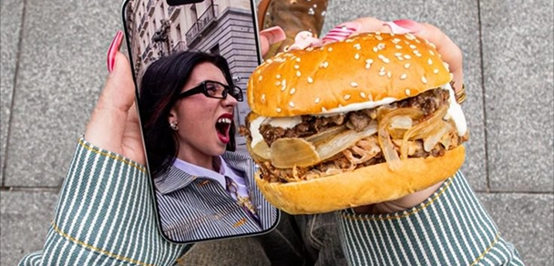 Person holding a smartphone in one hand showing an image of a woman with glasses and dark hair yelling or excited, and a large sesame seed hamburger with grilled onions in the other hand.
