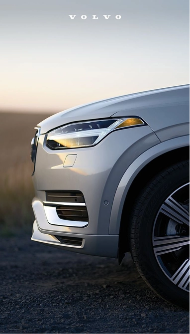 Close-up of the front side of a silver Volvo SUV showing the headlight, front grille, and part of the tire, with a blurred natural background at sunset.