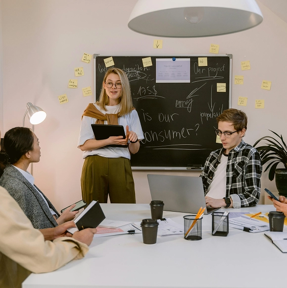 A young woman with glasses and a brown sweater draped over her shoulders stands holding a tablet, presenting to a group in a meeting room with a blackboard behind her covered in notes and sticky notes. Two people sit at the table, one with a laptop and the other holding a notebook, listening attentively. The table holds papers, coffee cups, and stationery in an informal collaborative work setting.