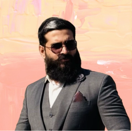 Man with a thick black beard and slicked-back hair wearing dark sunglasses and a gray three-piece suit with a white shirt and burgundy pocket square.