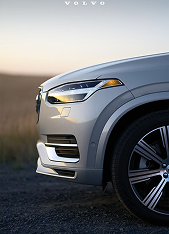 Front side view of a silver Volvo car focusing on the headlight, grille, and front wheel against a blurred outdoor background at sunset.