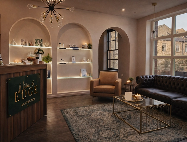 Cozy waiting room with warm lighting featuring a brown leather sofa, beige armchair, glass coffee table on a patterned rug, and wooden reception desk with 'Willow Edge Aesthetics' sign, softly lit shelves with decorative items, and large windows showing stone buildings outside.