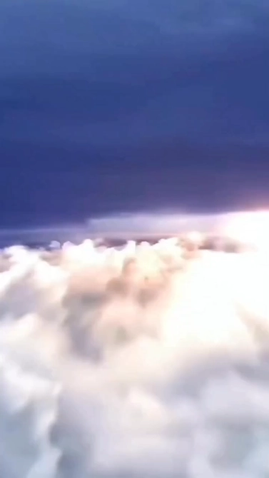 View above fluffy white clouds during a sunset or sunrise, with a dark blue sky above and soft light illuminating the clouds.