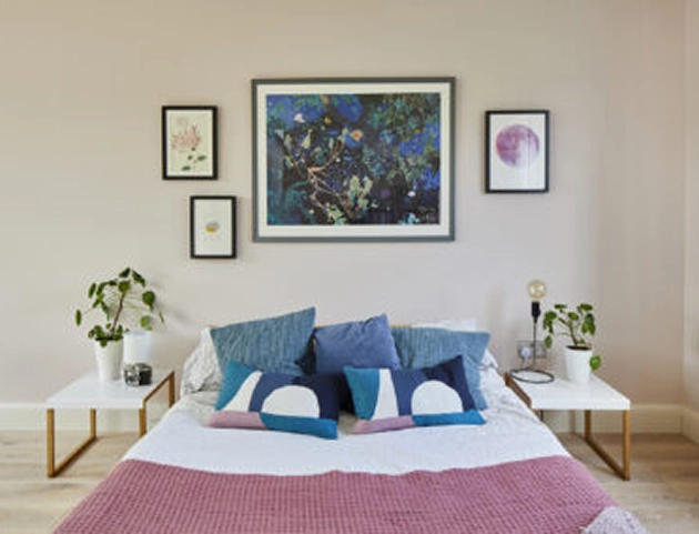 Bedroom with a neatly made bed featuring white sheets, a pink knitted blanket, and blue and navy decorative pillows, flanked by two white bedside tables each with a potted plant; three framed abstract art pieces hang on the beige wall above the bed.