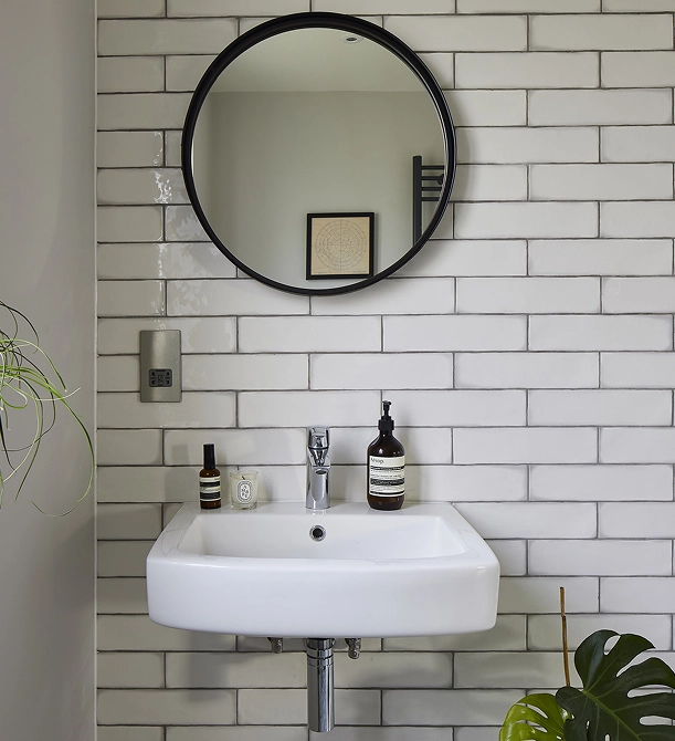Minimalist white bathroom sink mounted on a white tiled wall with rectangular tiles, a round black-framed mirror above it, a small potted plant in the lower right corner, and soap and skincare products on the sink.