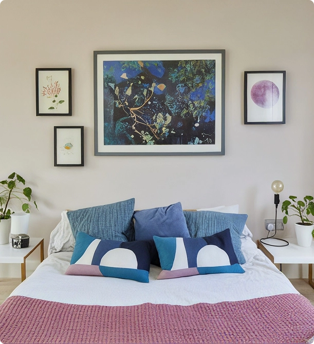 Bedroom with a bed featuring white sheets, a pinkish-purple textured blanket, and five decorative pillows in blue and patterned designs. Above the bed are four framed artworks: a large abstract nature-themed piece in the center, a purple circle on the right, and two smaller botanical-style prints on the left. Each side of the bed has a white bedside table; the left table has a plant and a small container, while the right table has a plant and a lamp with an exposed bulb.