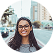 Smiling woman with long dark hair and glasses standing outdoors in an urban setting with buildings.