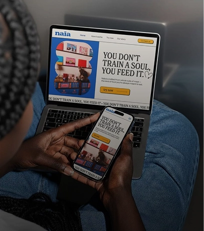 Person holding a smartphone displaying a webpage with the quote 'You don't train a soul, you feed it' while a laptop shows the same webpage with colorful shelves and a call-to-action button that says 'try now'.