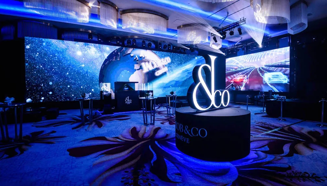 Dimly lit event space with large digital screens displaying a cosmic scene and racing cars, featuring a lit sign with '&co' on a round pedestal on a patterned carpet.