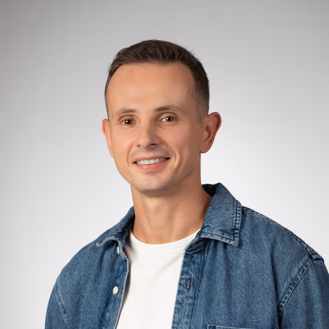 Smiling man with short brown hair wearing a denim jacket over a white shirt on a gray background. Portrait of Jacek Radziejewski.
