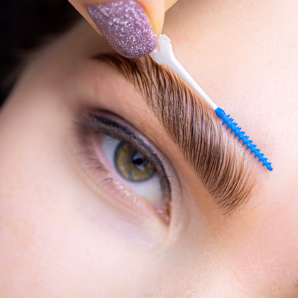 Close-up of a hand with glittery purple nail brushing a well-groomed eyebrow with a blue spoolie brush.