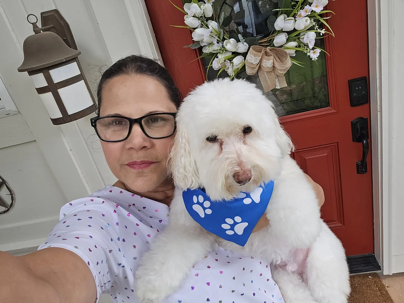 Woman wearing glasses holding a white fluffy dog with a blue paw-print bandana in front of a red door.