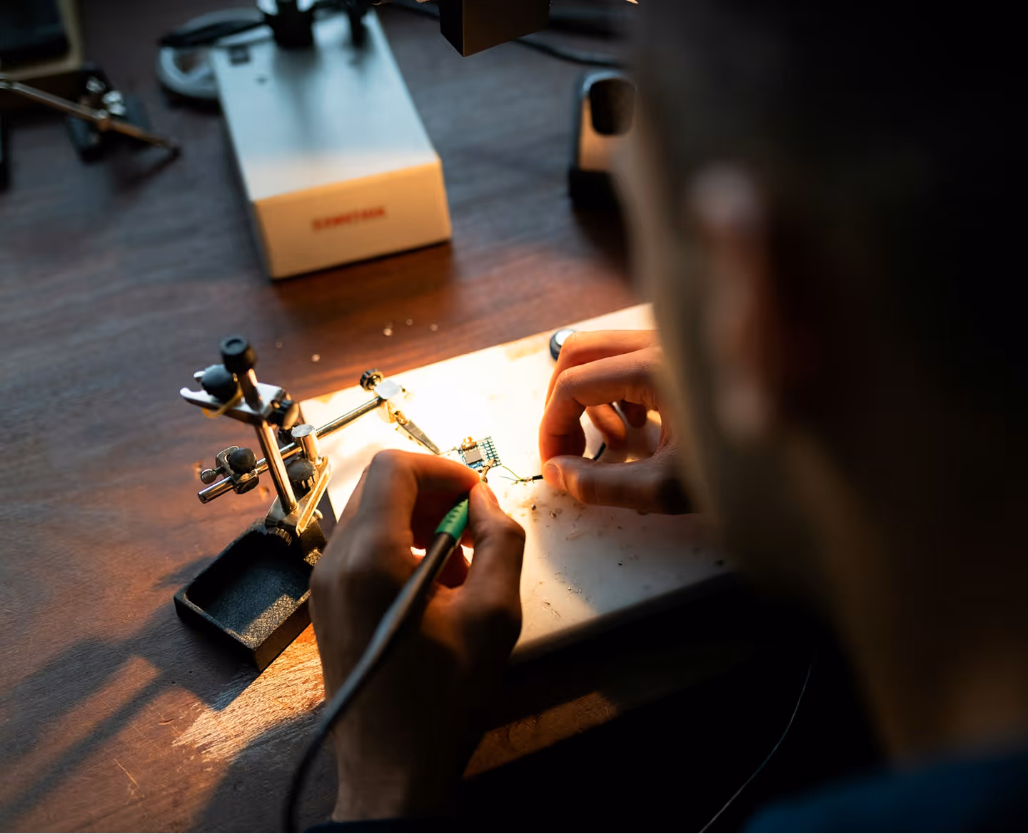Person soldering electronic components on a small circuit board at a wooden desk under warm light.