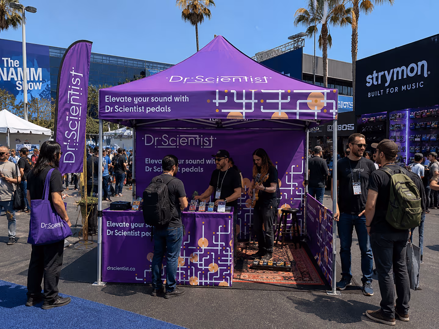 Outdoor Dr. Scientist booth with purple tent, displaying guitar pedals and people interacting at The NAMM Show.