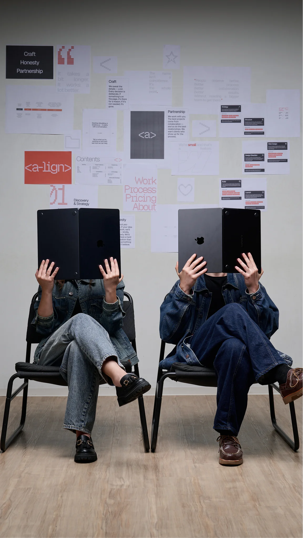 Tamara and Jan sitting on chairs reading laptops like newspapers with papers on wall behind them.