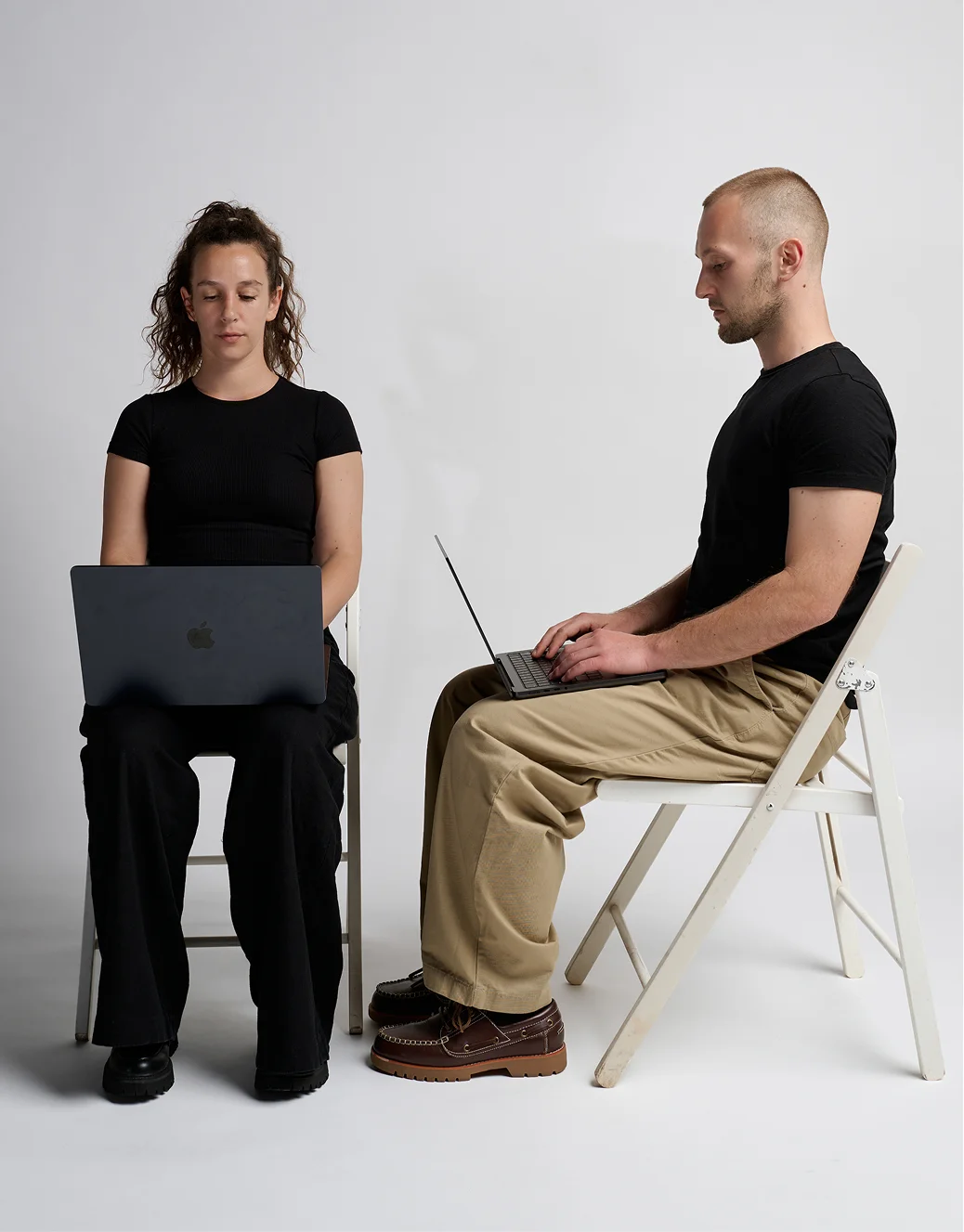 Tamara and Jan sitting on white folding chairs, working on laptops in a plain light room — representing the process.