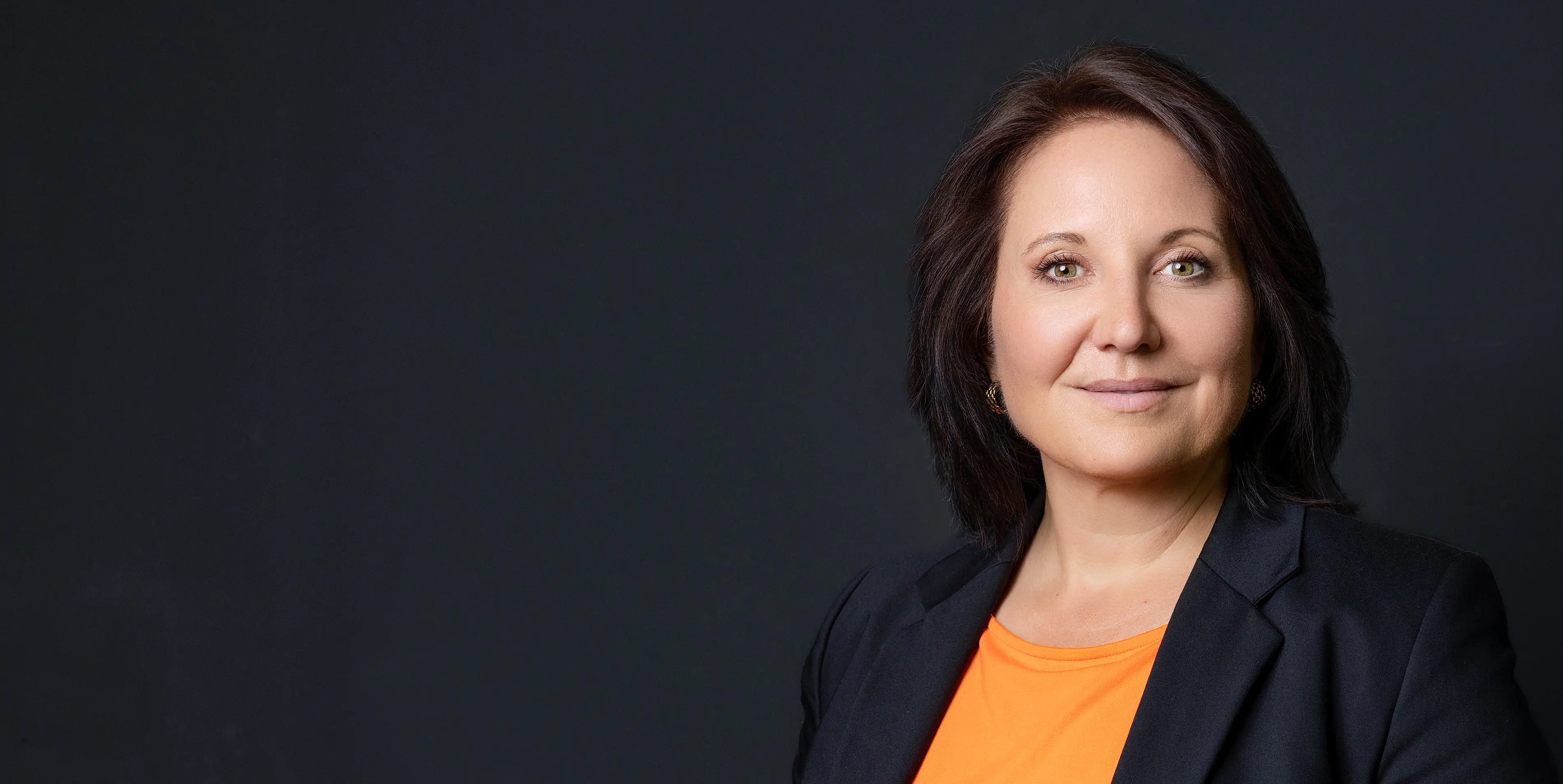 Marion Mossmann Schwarz im Portrait mit dunklem Blazer und orangefarbenem Oberteil vor dunklem Hintergrund.