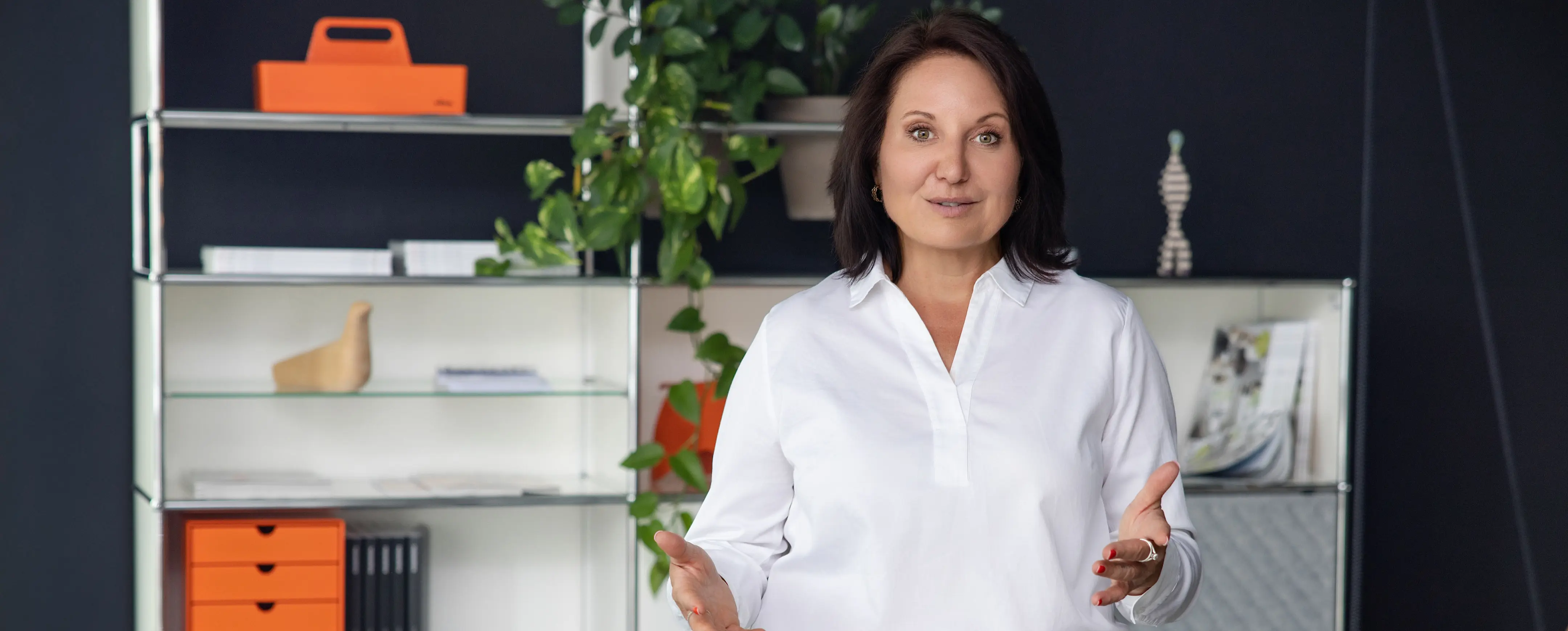Marion Mossmann Schwarz steht im weißen Hemd vor einem Regal in einem hellen Arbeitsraum.