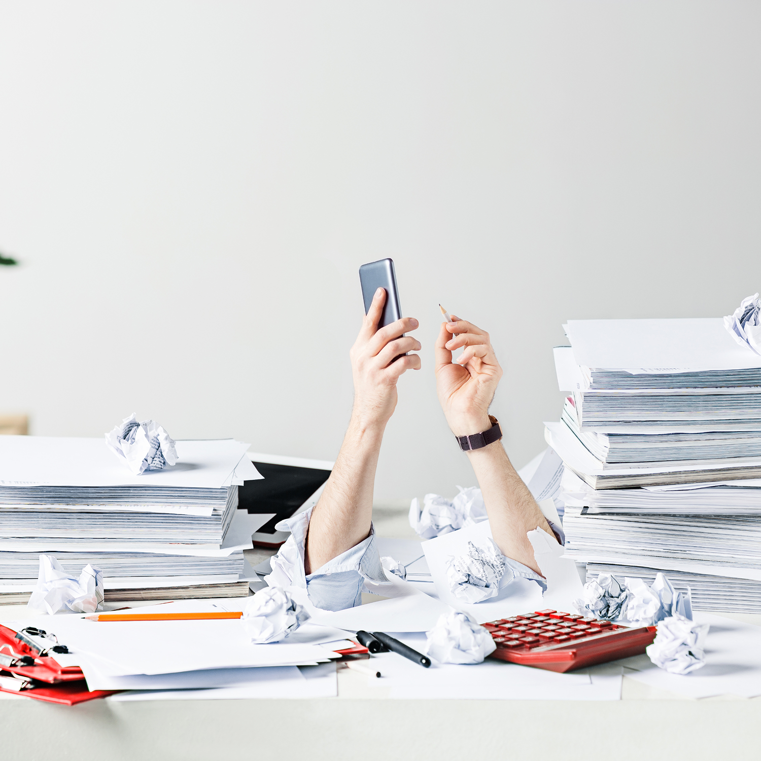 Person zwischen Papierstapeln am Schreibtisch mit Smartphone als Symbol für Überlastung und fehlende Zeit im Arbeitsalltag.