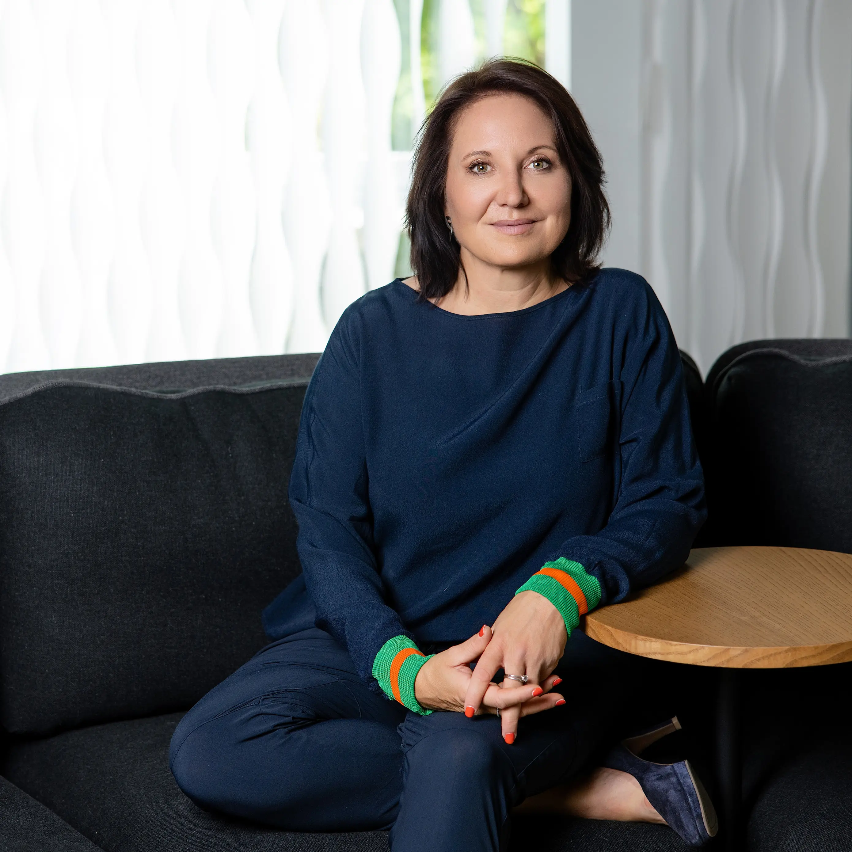 Marion Mossmann Schwarz sitzt in dunkelblauem Outfit auf einem Sofa neben einem runden Tisch.
