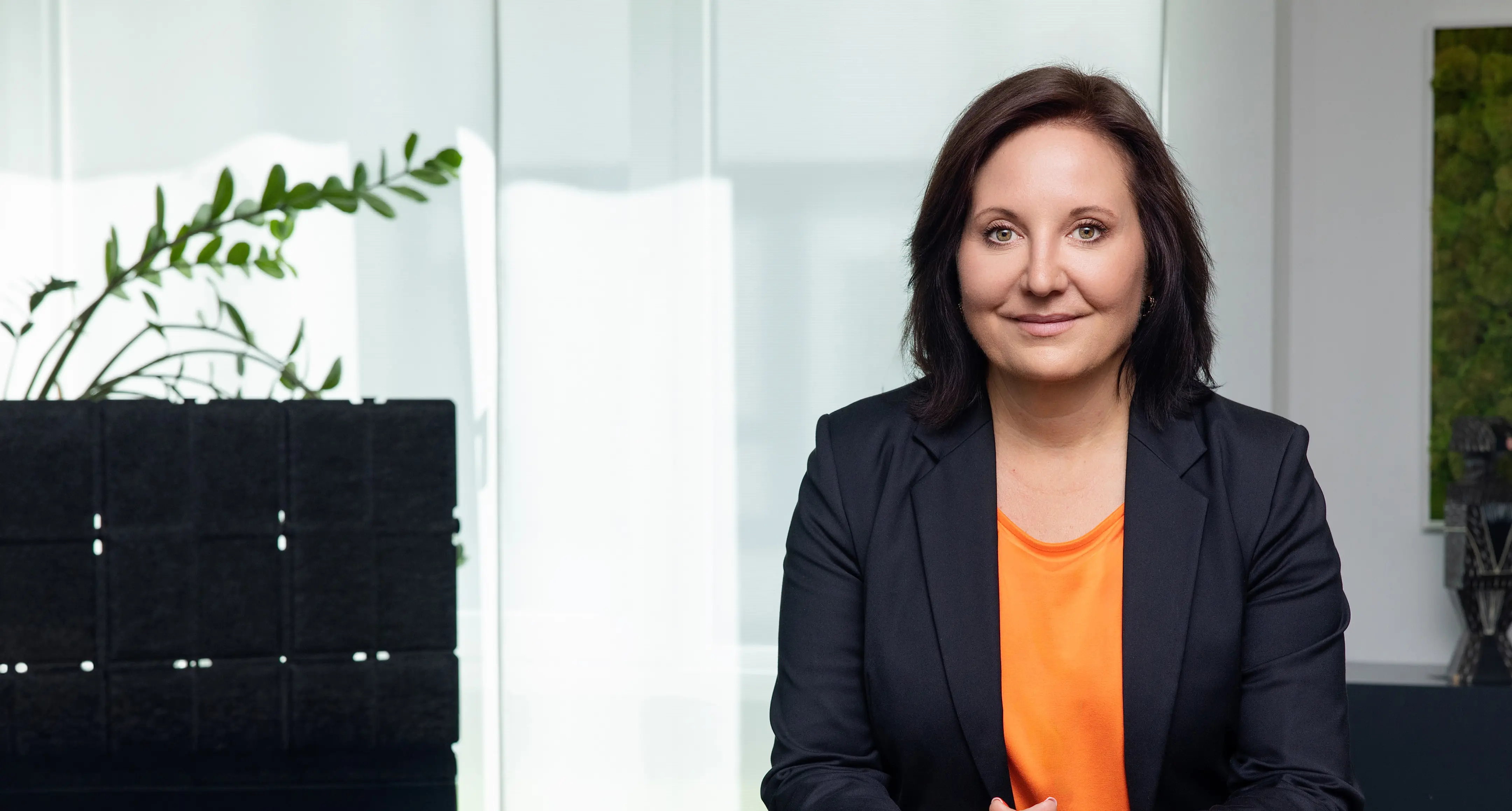 Marion Mossmann Schwarz sitzt im Blazer mit orangefarbenem Oberteil vor hellem Hintergrund.